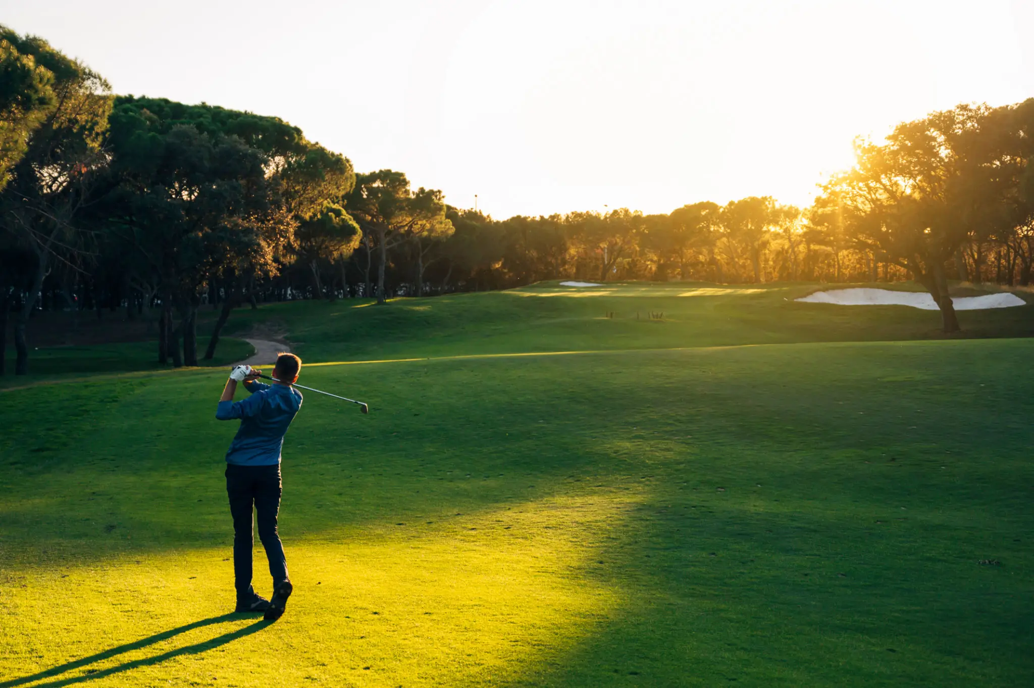 Golfer swinging on sunlit course at sunset.